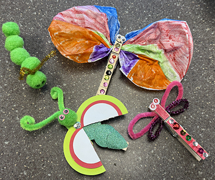 Bug crafts: a green pom-pom caterpillar, a colorful butterfly on a craft stick, a ladybug with green pipe cleaner antennae, and a clothespin dragonfly with pink pipe cleaner wings